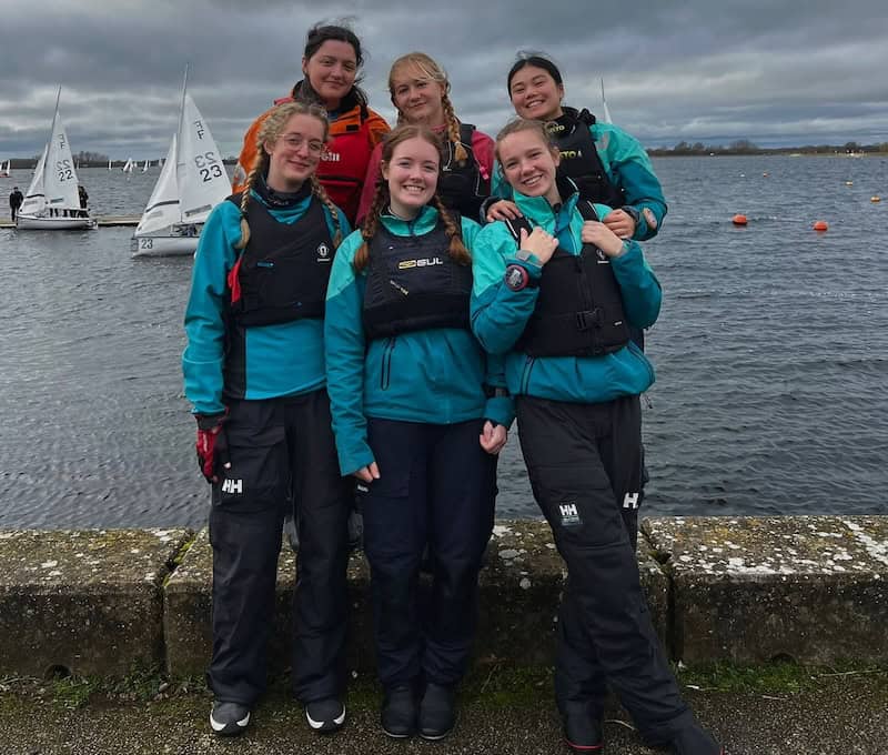The six members of Cambridge Women's team standing at the edge of Farmoor Reservoir with a couple of Firefly dinghies in the background